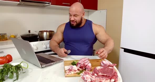 Cheerful Man Is Sitting at Home, Chopping Beef and Viewing Social Nets at Notebook