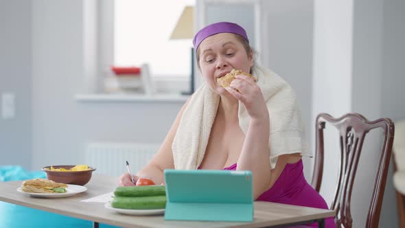 Young Plussize Woman Eating Sandwich Writing Calories Looking at Tablet Screen