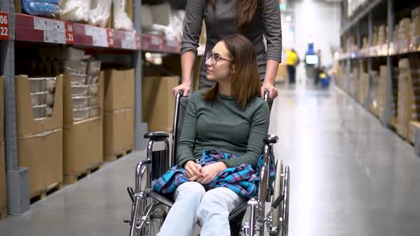 A Young Woman Is Sitting in a Wheelchair. Female Assistant Drives a Woman in a Wheelchair. Walking