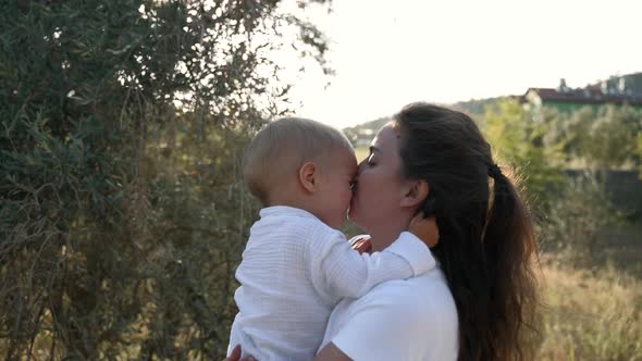 Young Mom Kisses Little Son Walking in Garden on Sunny Day