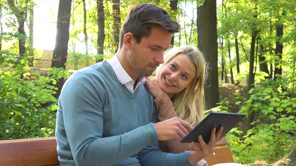 A Young Attractive Couple Looks at a Tablet on a Bench in a Park on a Sunny Day - Closeup