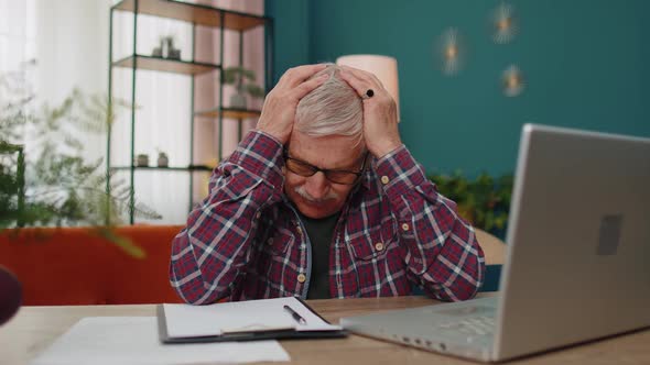 Senior Man Working on Laptop Computer Making Notes on Sheet of Paper Upset By Poor Bad Results