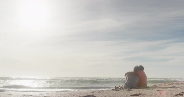 Hispanic senior couple sitting and embracing on beach at sunset