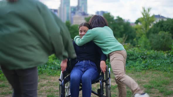 Beautiful Disabled Girl Smiling Sitting in Wheelchair Outdoors As Cheerful Friends Running Hugging