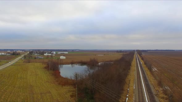 Cars, trucks and a bus traveling across an overpass running over railroad tracks next to a pond on a