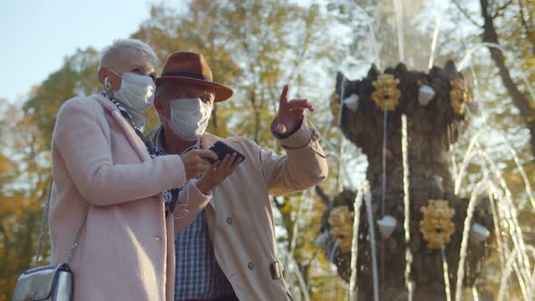 Senior Couple in Protective Mask Using Headphones and Smartphone Having Tour in City Park