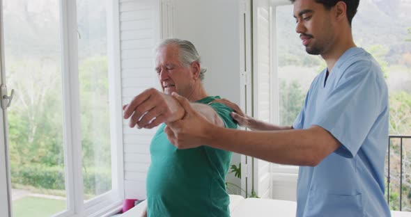 Biracial male physiotherapist slowing moving hand of senior man grimacing with pain at nursing home
