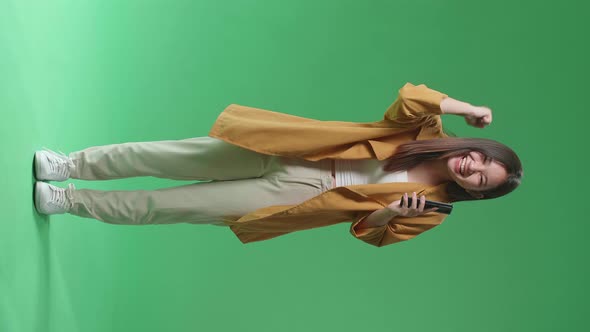 Full Body Of Young Asian Woman Celebrating With Mobile On Green Screen In The Studio