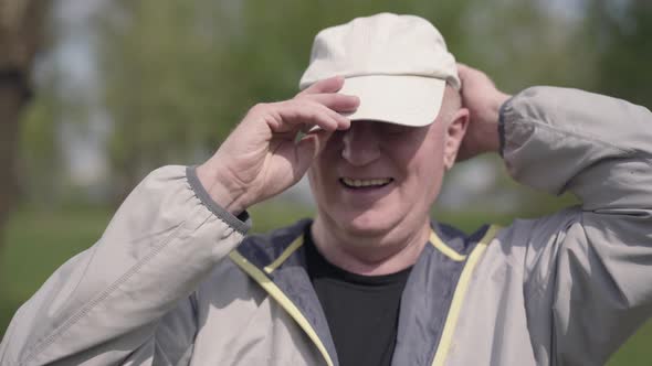 Positive Mature Man Putting the Cap on the Head in the Park, Looking in the Camera