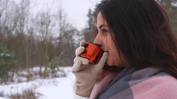 Young Cute Swarthy Brunette Woman Drinking Hot Drink From Orange Thermos Cup in Snowy Winter Forest