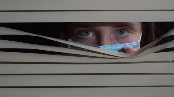 Girl in protective mask looking out the window through the blinds to street