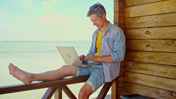 Portrait Optimistic Smiling Man Freelancer Working Outdoors on Beach By Blue Sea Drinking Coffee