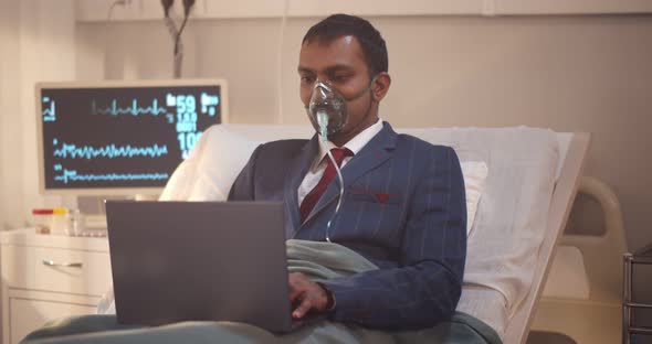 Indian Businessman with Oxygen Mask in Suit Working with Laptop Lying on Hospital Bed