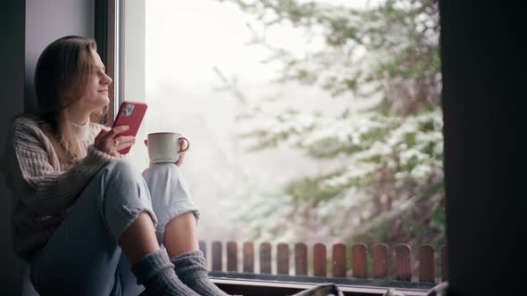 A Young Girl Sits on the Windowsill and Drinks Coffee While Using Her Smartphone