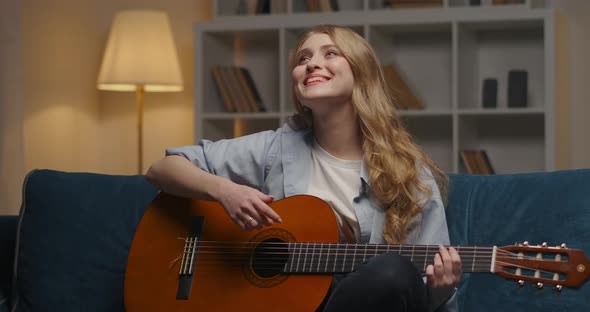 Cheerful Young Lady with Guitar Is Having Fun at Home at Evening Sitting Alone in Cozy Apartment and