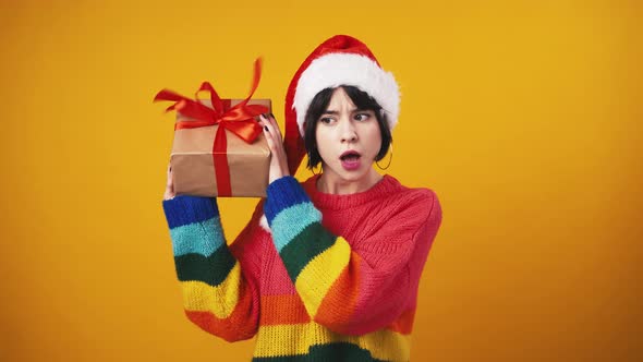 Young Woman in Santa Hat Catching Gift Box and Shaking It Guessing What's Inside Orange Background