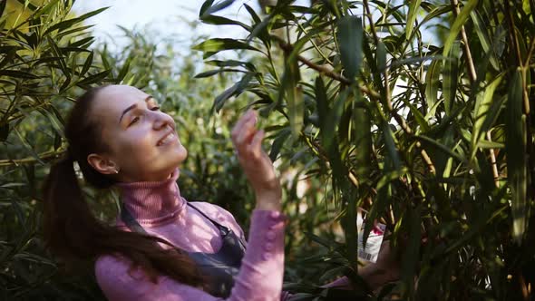 Happy Smiling Female Gardener in Apron Examining Leaves of Different Trees While Walking Among Rows