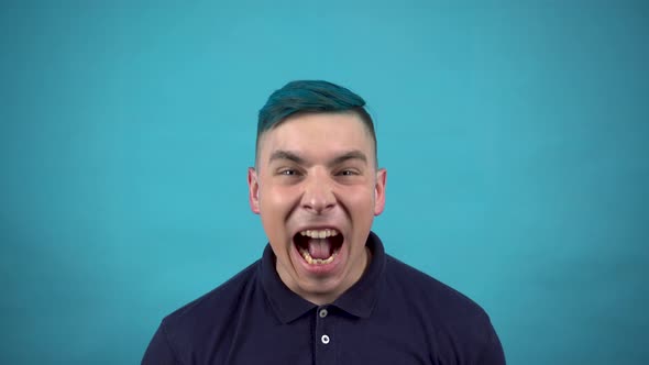 A Young Man in Glasses with Blue Hair Screams in Anger. Emotional Man Screaming on a Blue Background