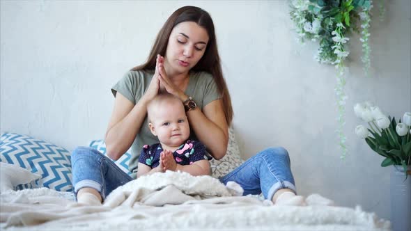 Young Darkhaired Mother and Infant Girl are Sitting on a Bed and Clapping