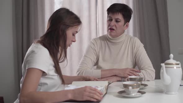 Senior Caucasian Woman Watching Photo Album with Adult Daughter at Home. Sad Lady Telling Young Girl