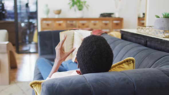 Happy african american woman lying on sofa in living room, reading book