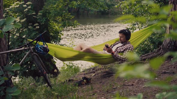 Man Travels on Bicycle Relaxing in Green Hammock Surfing Internet on Smartphone Listening Music on