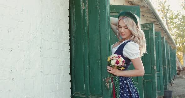Blonde in Ethnic Costume with Bouquet of Flowers Stands in Pose at Stable