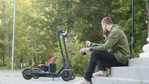 Couple Man and Woman are Sitting on Stairs in Park and Drinking Coffee There are Electric Scooters