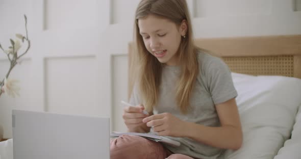 Teen Girl Is Preparing To Online Lesson By Internet, Holding Copybook, Sitting on Bed in Front of