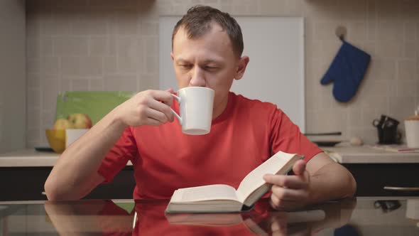 The guy is drinking tea and reading a book while sitting in the kitchen on a bright sunny day