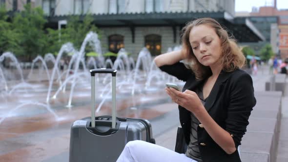 Young woman with suitcase on plaza with jet fountains at front of the train station.