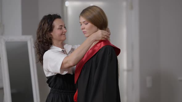 Side View Happy Loving Mature Mother Putting Graduation Gown on Charming Daughter