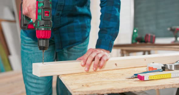 Concentrated Young Bearded Carpenter Wearing Jeans and a Plaid Flannel Shirt Drilling a Hole in a