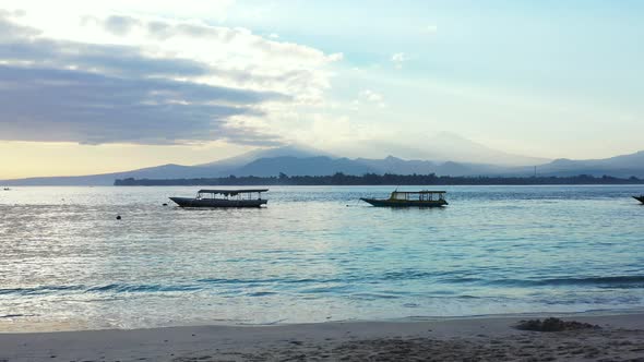 Long boats floating in the peaceful sea, sandy beach and hills in the background covered in the clou