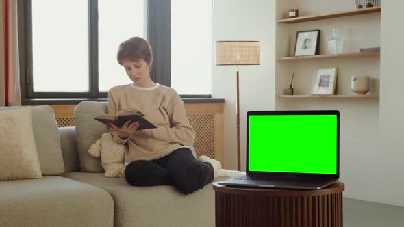 A Laptop with a Green Screen Stands on a Table Against of a Woman Reading a Book