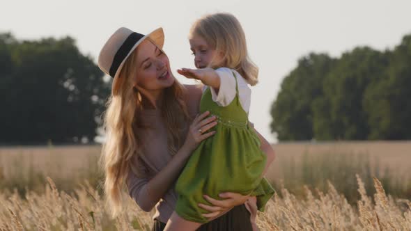 Young Woman Mother Caucasian Mommy in Hat Stands in Wheat Field Holding Small Girl Daughter Child