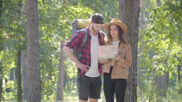 Young Couple of Caucasian Hikers Walking in Sunny Forest, Looking at Paper Map, and Leaving