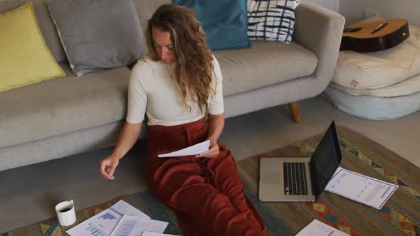 Caucasian woman working in living room, sitting on floor with laptop and paperwork, drinking coffee