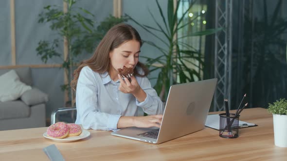 Young Businesswoman Eating a Glazed Donut While Working on Laptop in Office