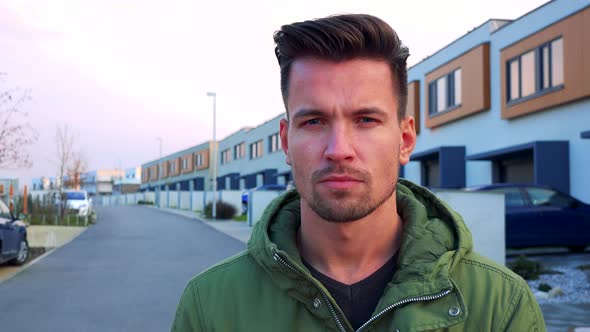 A Young, Handsome Man Stands on a Road, Houses in the Background and Looks at the Camera