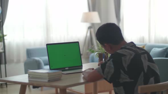 Teenage Boy Sitting At His Desk Learning Online On A Laptop With Green Mock-Up Screen At Home