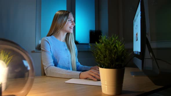 Smiling Woman Working on Computer at Night. Smiling Female in Checkered Shirt Sitting at Lit By