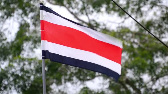Close-up shot of Costa Rican Flag waving in the wind in front of green tree tops in background. Clou