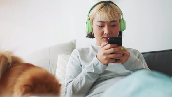 Cheerful Asian woman looking at phone and stroking her dog on the bed