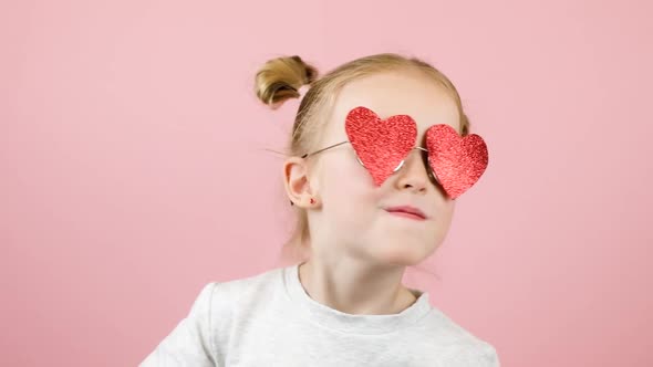 Funny Little Blonde Girl Smiling and Dancing in Red Heart Shape Sunglasses on Pink Background