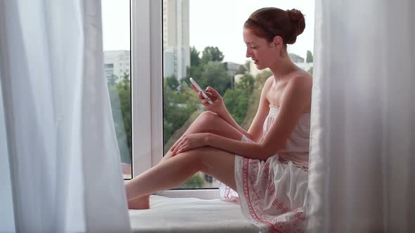 Young Beautiful Woman with a Smartphone on the Windowsill