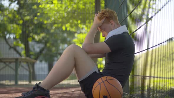 Side View of Exhausted Basketball Player Sitting at Mesh Fence Holding Head in Hands. Portrait of