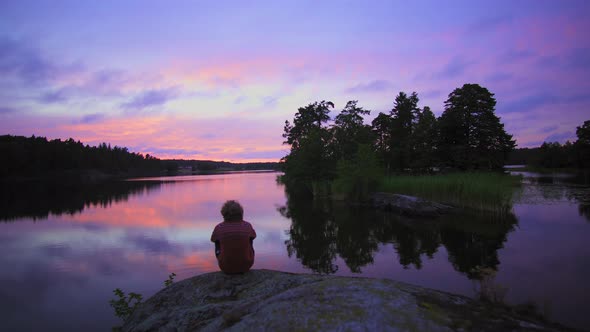 Walking towards a man sitting at a rocky shore, of a lake, a purple sky, at a colorful sunset or dus