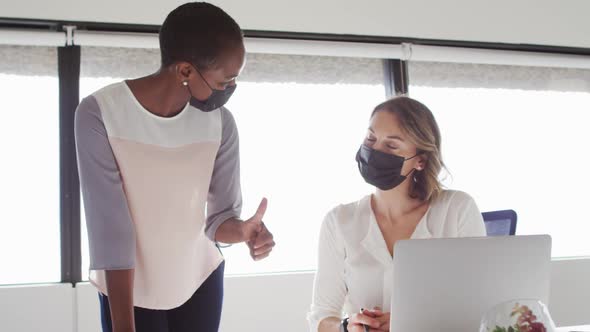 Two diverse female colleagues wearing face masks looking at laptop and discussing in office
