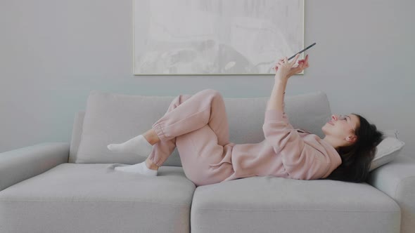 Young woman lying and resting on sofa, tapping on tablet, reading news and browsing social networks.